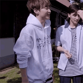 two young men are standing next to each other in front of a building . one of the men is wearing a white hoodie .