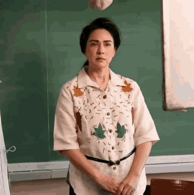 a woman is standing in front of a green board in a classroom wearing a shirt with leaves on it .