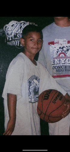 a young boy holding a wilson basketball in front of a man wearing a vince carter shirt
