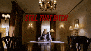 a man sits at a table in a room with the words still that bitch above him