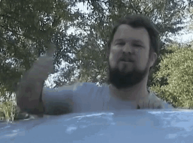 a man with a beard is giving a thumbs up in front of trees .