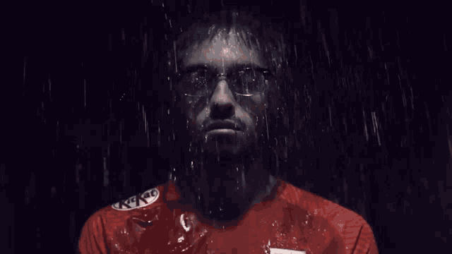 a man wearing glasses and a red shirt is standing in the rain with water coming out of his face