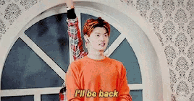 a young man in an orange sweater is standing in front of a window and says `` i 'll be back '' .