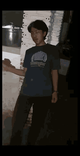 a young man wearing a wrangler t-shirt stands in a dark room