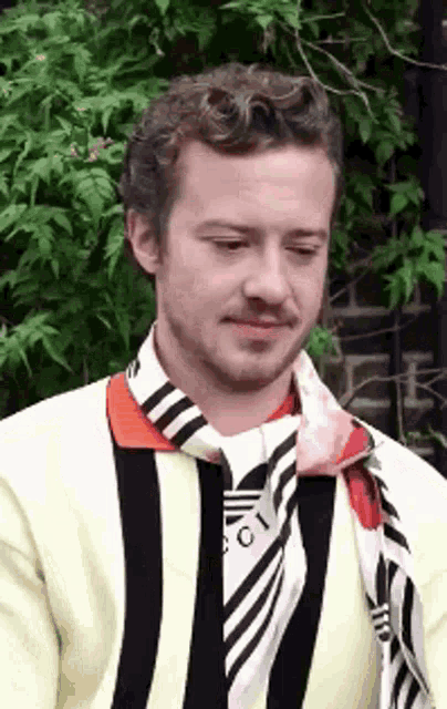 a man wearing a scarf around his neck is standing in front of a tree