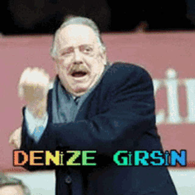 a man in a suit and scarf stands in front of a sign that says " denize girsin "