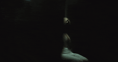 a woman in a white dress sits in a dark room with a circle on the wall above her