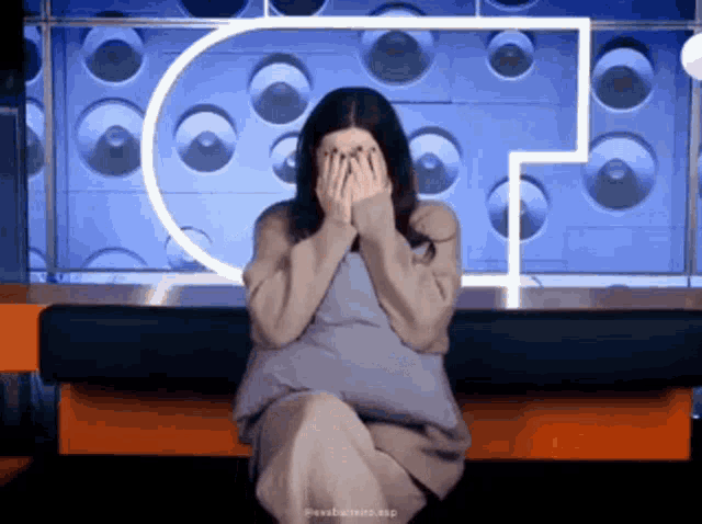 a woman is covering her face with her hands while sitting in front of a wall with circles on it .