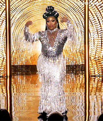 a drag queen is standing on a stage wearing a long silver dress with fringe .