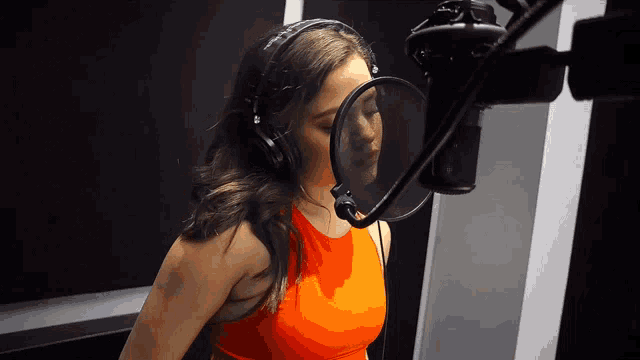 a woman wearing headphones and an orange top sings into a microphone