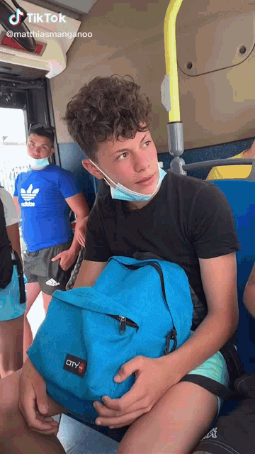 a boy wearing a face mask is sitting on a bus holding a blue city backpack
