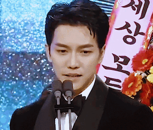 a man in a tuxedo is speaking into a microphone with a sign in the background that says ' a '