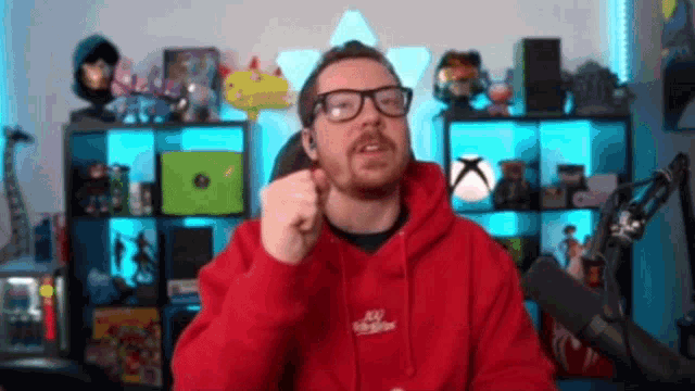 a man wearing glasses and a red hoodie is giving a thumbs up in front of a microphone .
