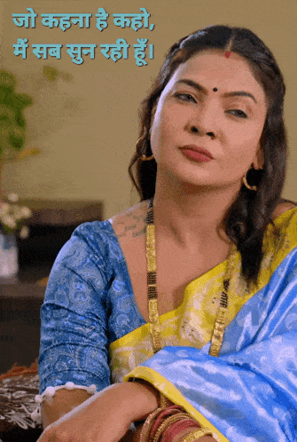 a woman in a blue and yellow saree is sitting at a table in front of a wall that says jo kahana he kaho