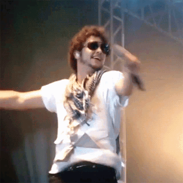 a man wearing sunglasses and a scarf is singing into a microphone on stage