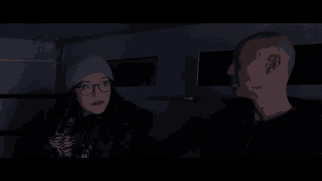 a woman wearing glasses and a beanie talks to a bald man in a dark room
