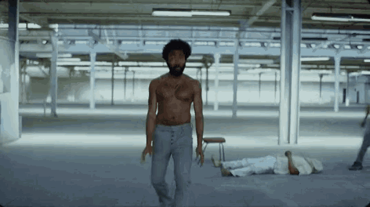 a man without a shirt is walking in an empty room while a man laying on the floor .