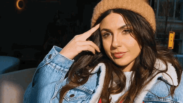 a woman wearing a denim jacket and a beanie holds her hand to her head