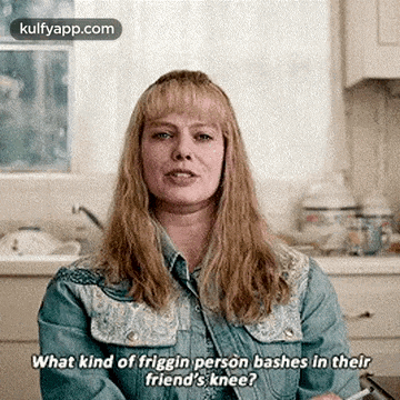 a woman in a denim jacket is sitting at a table in a kitchen and talking to someone .