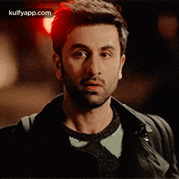a man with a beard is wearing a leather jacket and a sweater and looking at the camera .