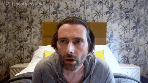 a man with a beard is sitting in front of a bed with the words @itsscottiesstark above him