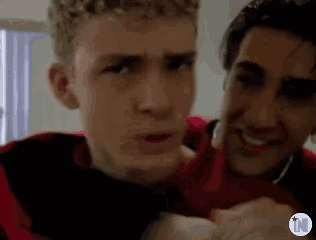 two young men are standing next to each other in a room . one of the men is wearing a red shirt .