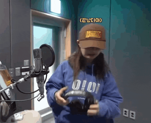a woman wearing a blue hoodie and a brown hat holds a pair of headphones in front of a microphone