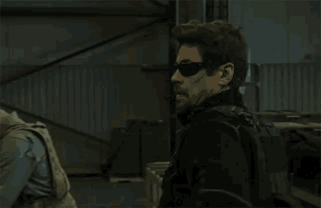 a man wearing sunglasses stands in a dark room next to another man