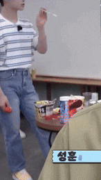 a man in a striped shirt and jeans is standing in front of a table holding a red object .