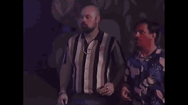two men are standing next to each other in a dark room and one of them is smoking a cigarette .