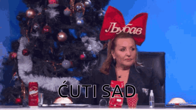 a woman with a red bow on her head is sitting in front of a christmas tree and says cuti sad