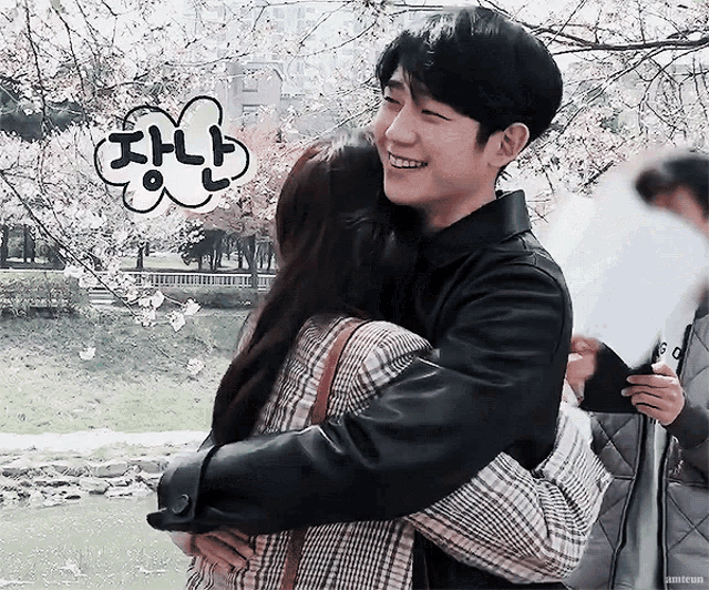a man in a black jacket is hugging a woman in front of cherry blossom trees