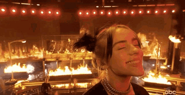 billie eilish is smiling while standing in front of a stage full of flames .