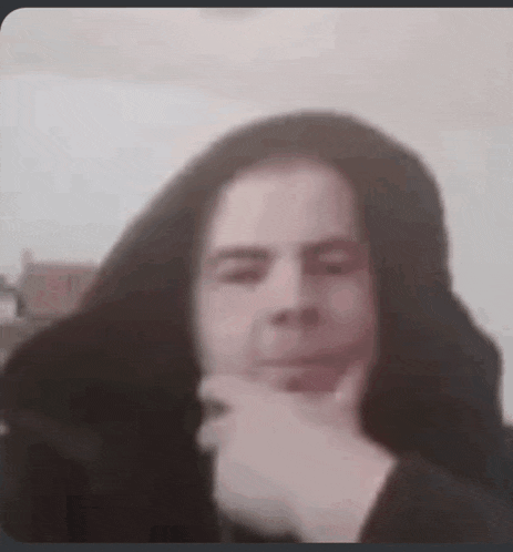 a man with long hair is holding his hand to his chin and looking at the camera .