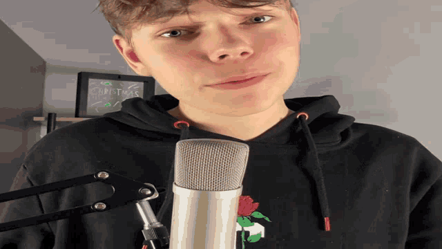 a young man stands in front of a microphone wearing a black hoodie that says christmas on it