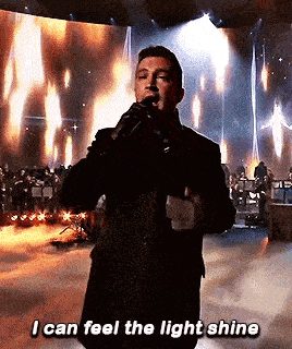 a man is singing into a microphone on a stage while saying `` i can feel the light shine '' .