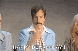 a man with a mustache is sitting in front of a group of people and saying happy burrfday .