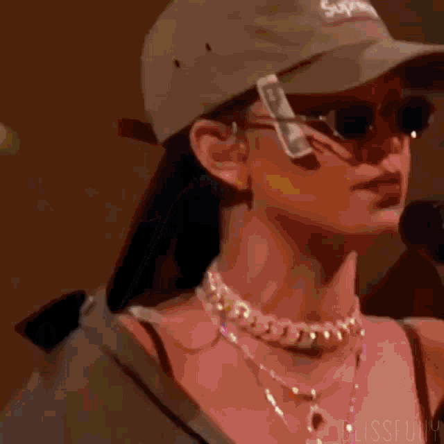a woman wearing a hat , sunglasses , and a necklace is standing in front of a microphone .