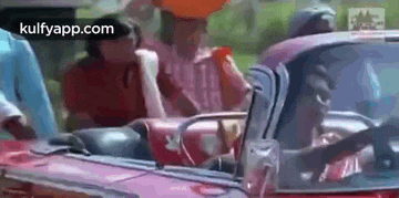 a man is driving a red car with a group of people sitting in the back seat .