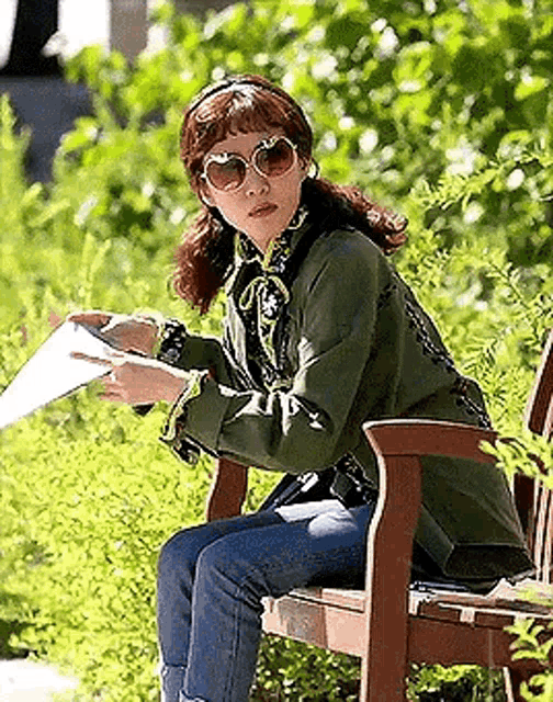 a woman wearing sunglasses is sitting on a park bench holding a piece of paper .