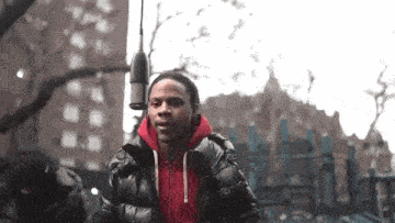 a man in a red hoodie is standing in front of a microphone in a park .