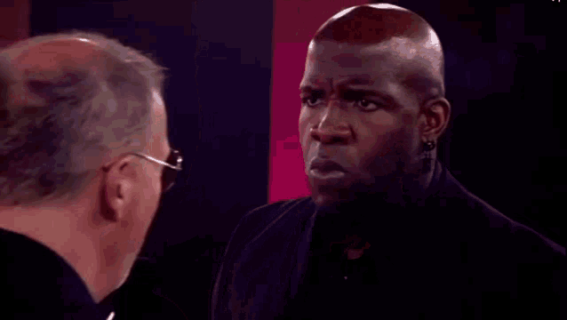 two men are looking at each other in a dark room and one of them is wearing earrings .