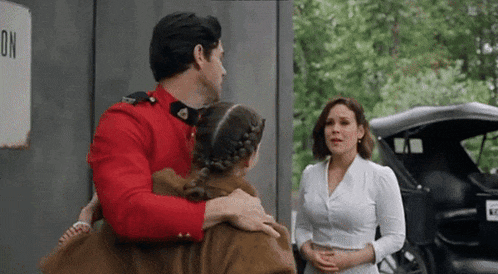 a man in a red uniform is holding a little girl in his arms while a woman looks on .