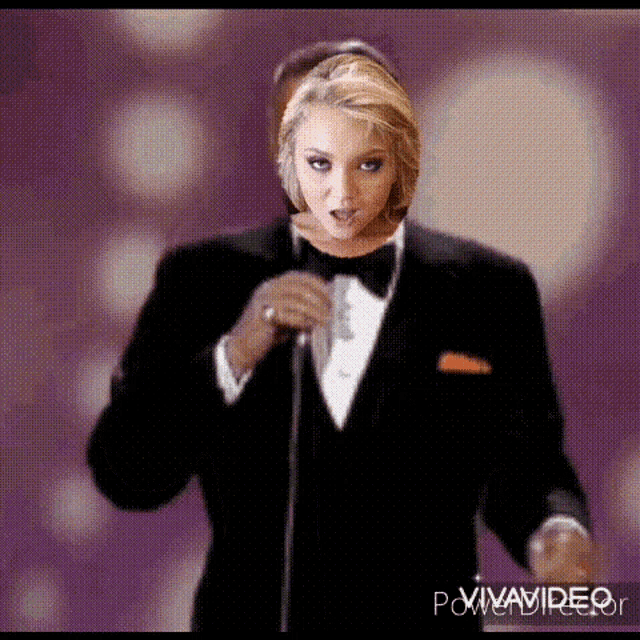 a woman in a tuxedo is singing into a microphone while a man looks on