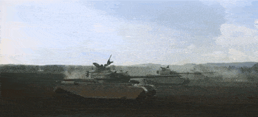 two military tanks are driving through a field .