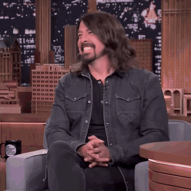 a man with long hair is sitting in a chair with his hands folded and smiling