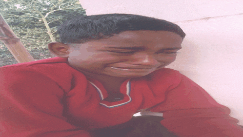 a young boy in a red shirt is crying while leaning against a wall