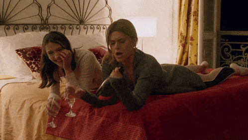 two women are laying on a bed and one is holding a glass of wine