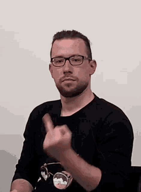 the man is wearing glasses and a black shirt and is giving the middle finger .