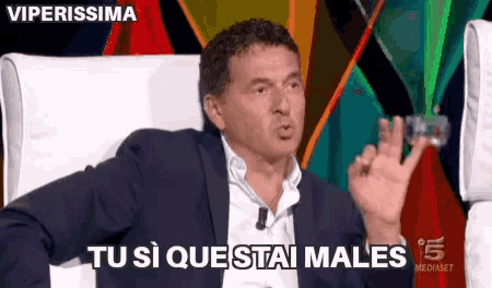 a man in a suit is sitting in a chair and saying " tu si que stai males " .